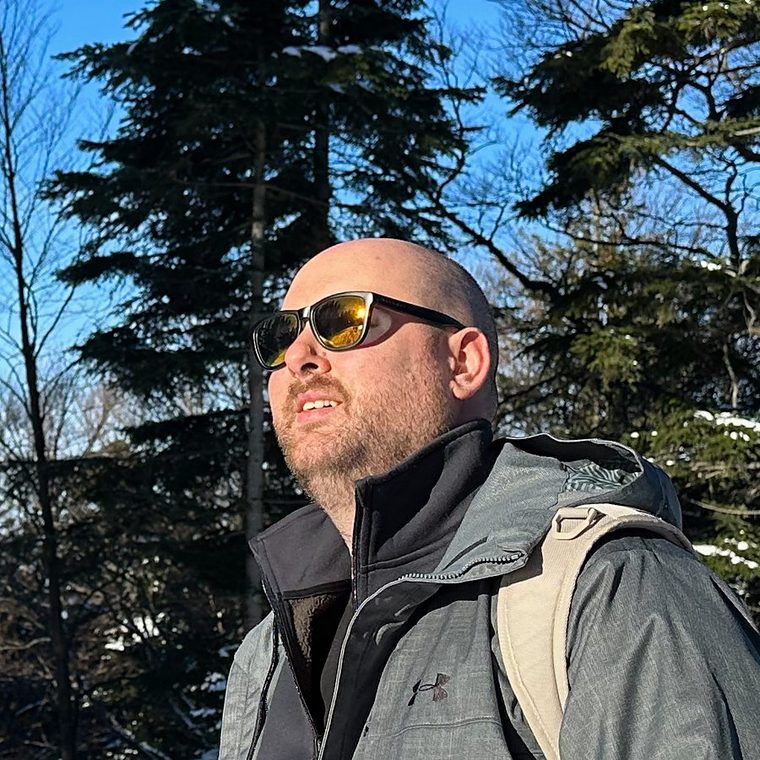 Tilen outdoors in winter sunlight, wearing sunglasses, with snow-covered pines behind him.